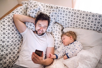 father with smartphone relaxes beside sleeping son